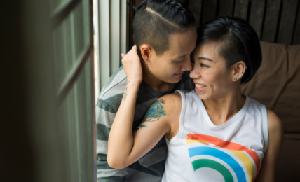 Two women in a romantic embrace. one woman is wearing a tank top with a rainbow on it.